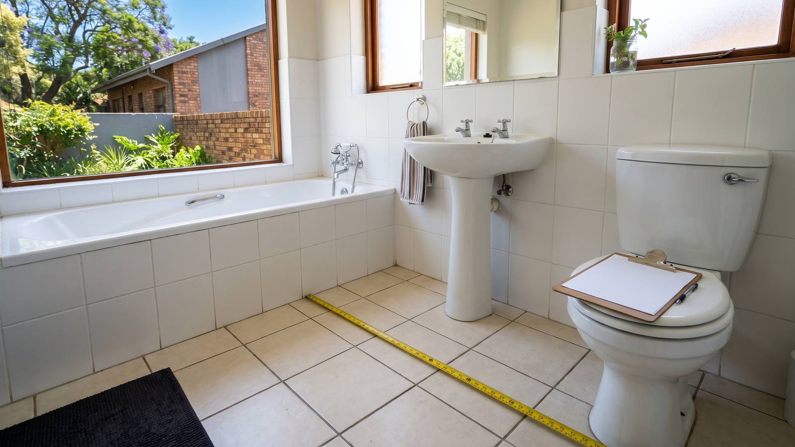 Tape measure stretched across a bathroom floor beside a bath and basin while a renovation quote is measured up, clipboard on the closed toilet lid.